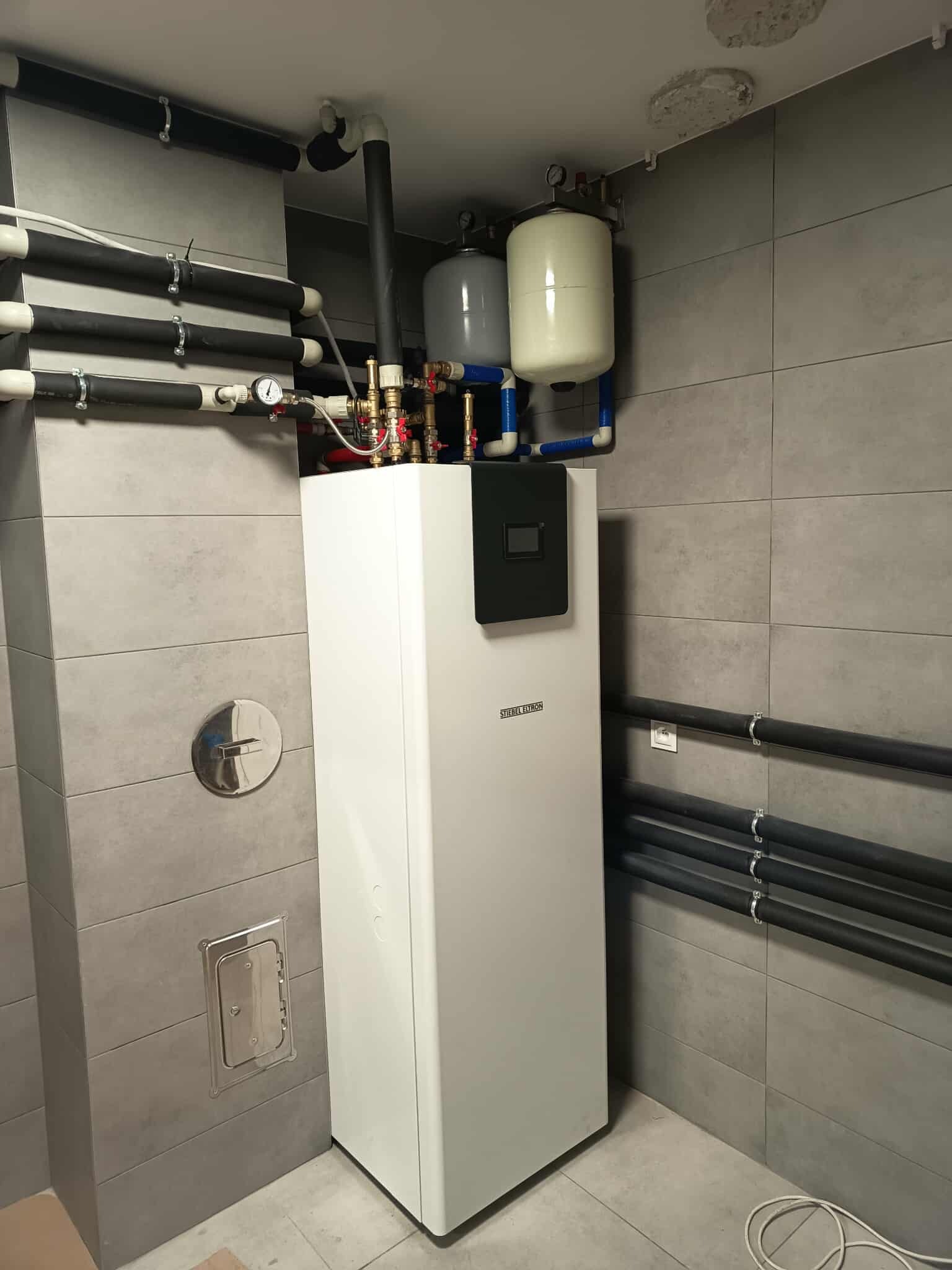 Biała pompa ciepła Stiebel Eltron w nowoczesnej kotłowni z szarymi płytkami, widoczne rury instalacji i zbiorniki wyrównawcze. Całość tworzy schludny i funkcjonalny system grzewczy.