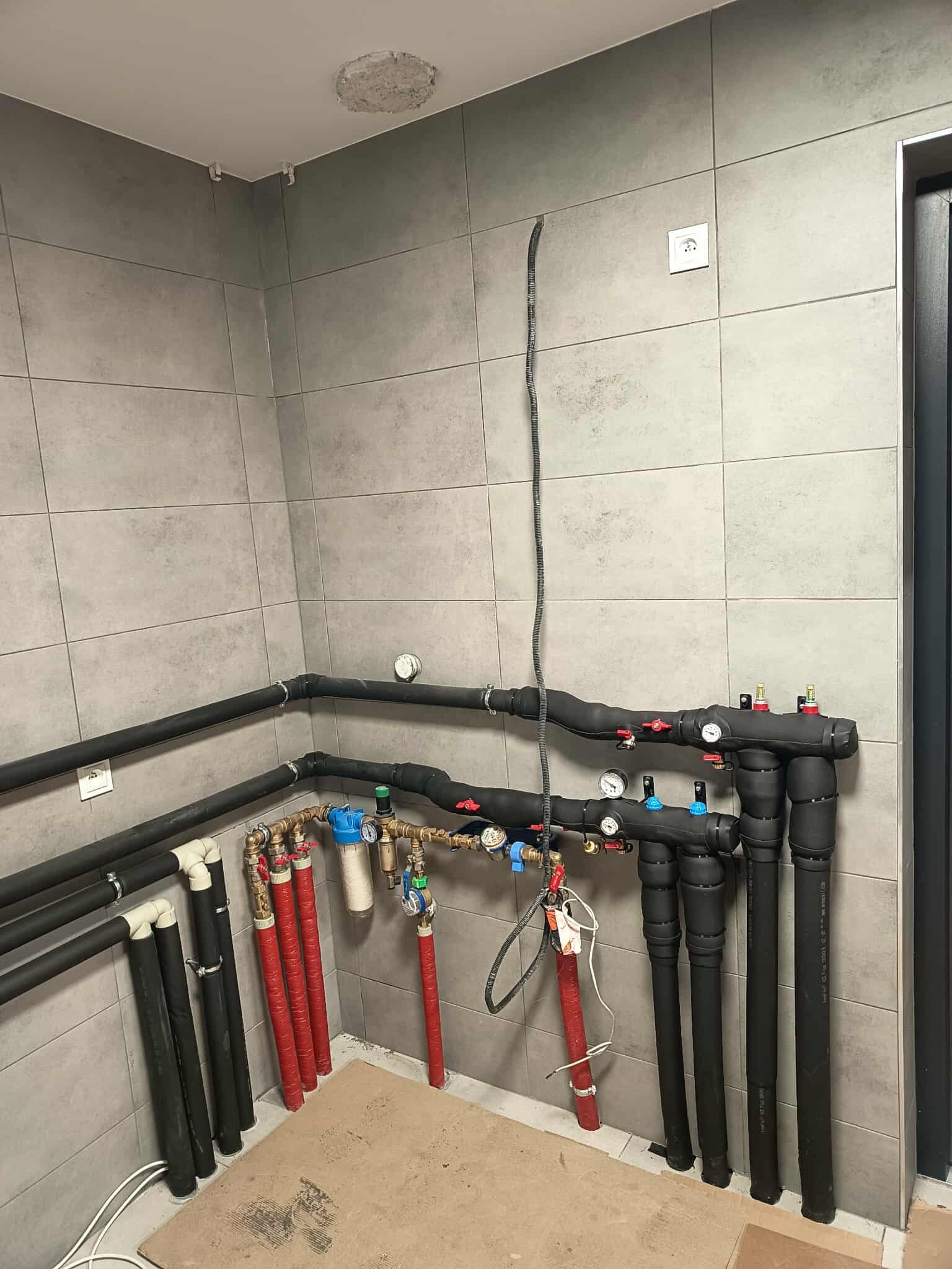 Złożona instalacja grzewcza z rurami w izolacji, manometrami i zaworami, zamontowana na szarej, kafelkowanej ścianie w pomieszczeniu technicznym. Widoczne elementy miedziane i plastikowe.