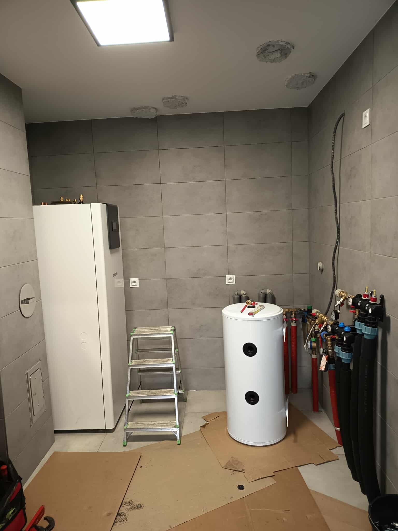 Biała pompa ciepła i zbiornik buforowy w szarej łazience z płytkami, widoczne rury i drabina. Montaż instalacji grzewczej w trakcie prac wykończeniowych.