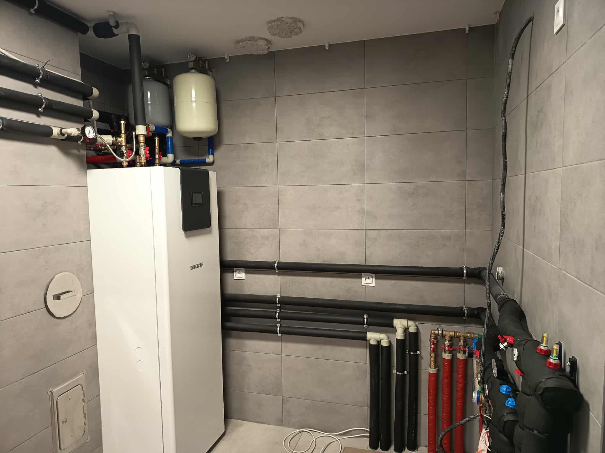 Biała pompa ciepła Stiebel Eltron w pomieszczeniu z szarymi płytkami. Widoczne rury, zbiorniki wyrównawcze i elementy instalacji grzewczej. Nowoczesna instalacja w kotłowni.