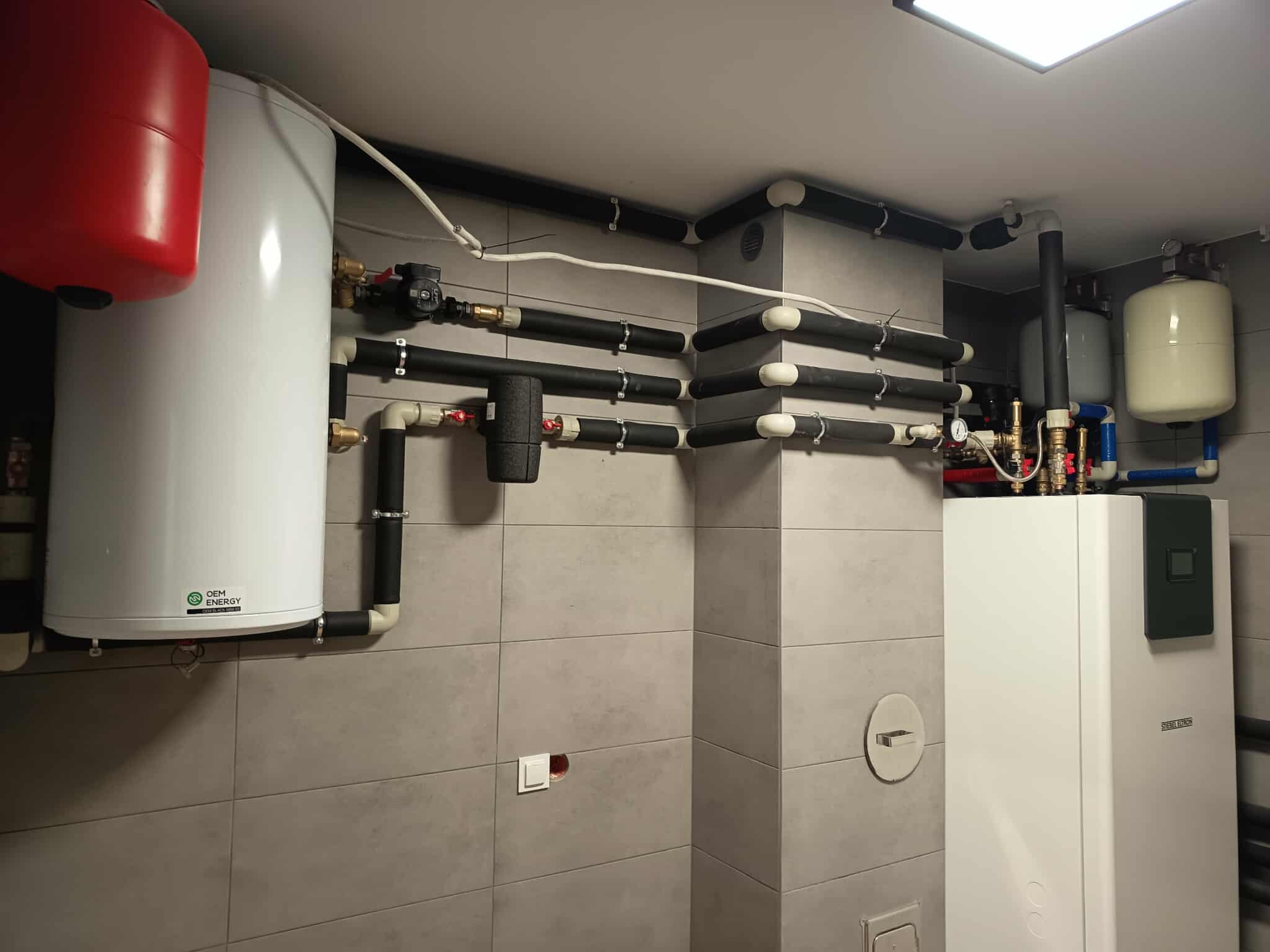Złożona instalacja grzewcza z rurami i zbiornikami w kotłowni, widok z dołu. Realizacja w Białobrzegi. Szare płytki na ścianach.