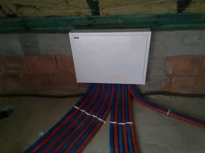 Rozdzielacz ogrzewania podłogowego w białej szafce, z widocznymi rurami w kolorze niebiesko-czerwonym, zamontowany na ścianie z cegieł i betonu.