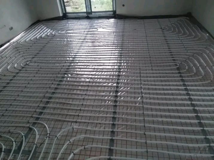 Ułożone rury ogrzewania podłogowego na siatce, widok z góry, przed wylaniem posadzki. Białe rury tworzą regularny wzór na czarnej siatce.