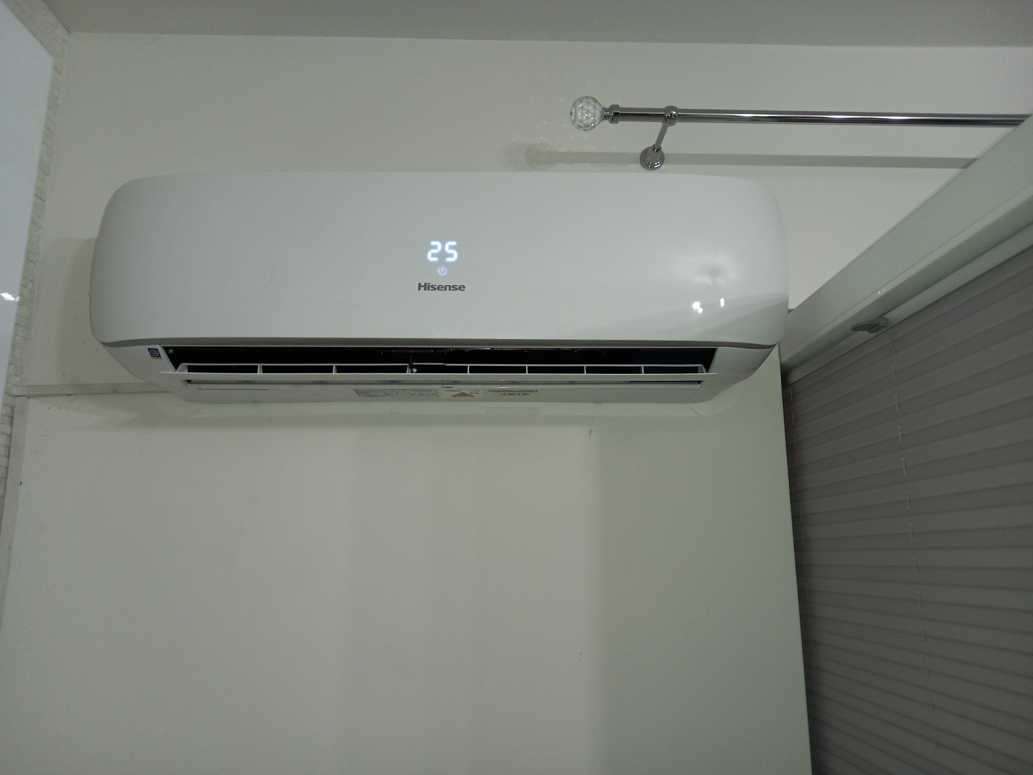 Biały klimatyzator Hisense zamontowany na jasnej ścianie, wyświetlający temperaturę 25 stopni Celsjusza, z widoczną karniszą z ozdobną końcówką i fragmentem żaluzji w tle.
