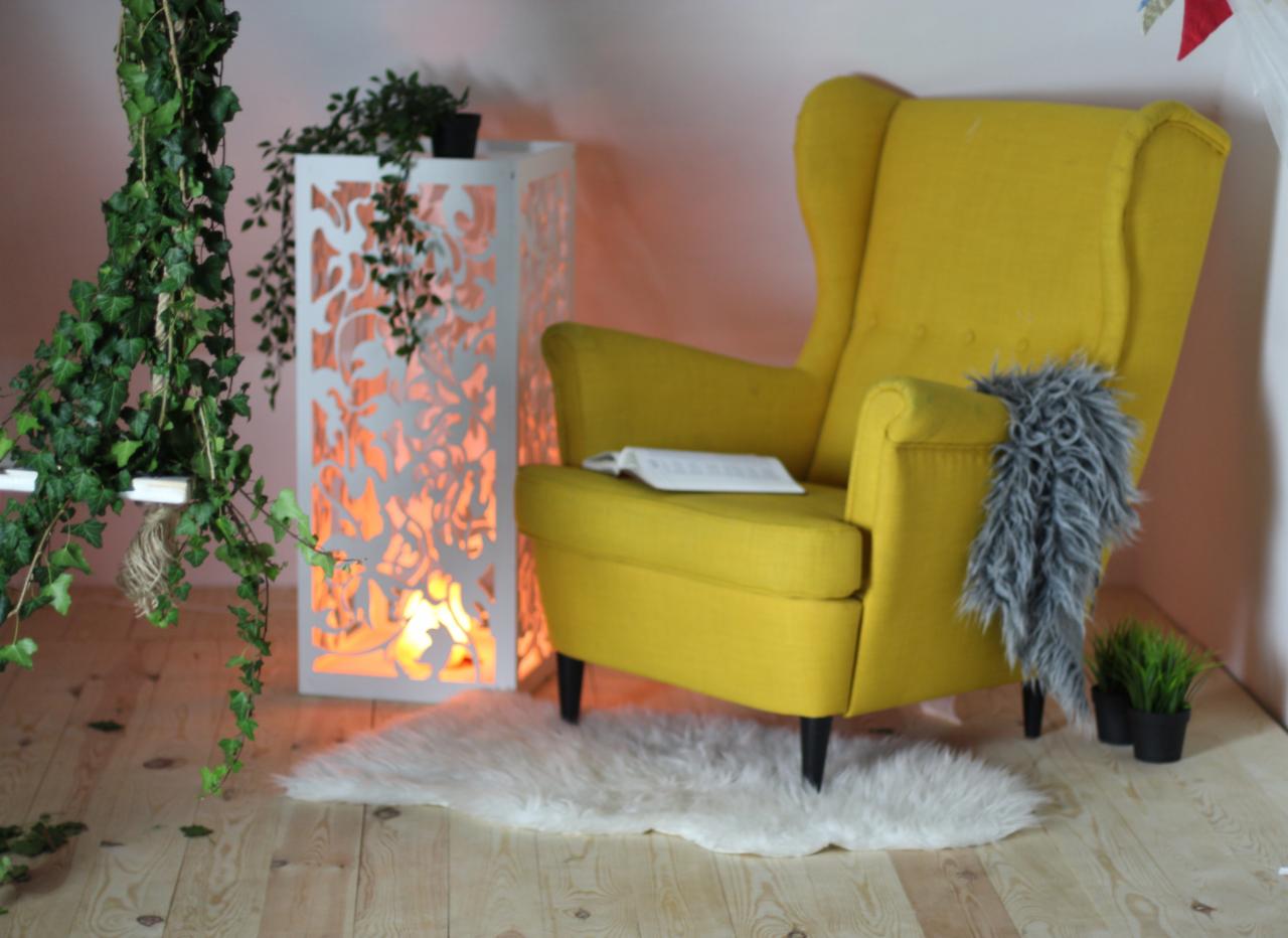 Żółty fotel uszak z książką i szarym pledem, biały lampion z ażurowym wzorem, pnącza bluszczu i dywanik z długiego włosia na drewnianej podłodze tworzą przytulną aranżację wnętrza.