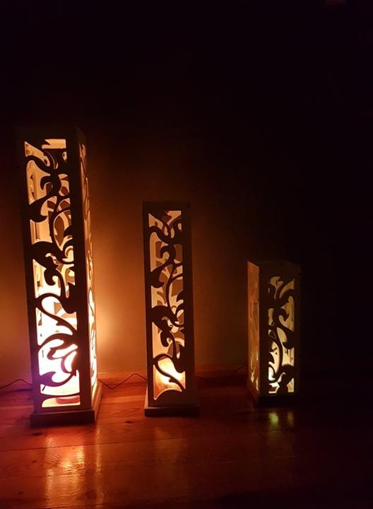 Trzy drewniane lampy podłogowe z wycinanymi wzorami roślinnymi, o różnej wysokości, ustawione na drewnianej podłodze w ciemnym pomieszczeniu.