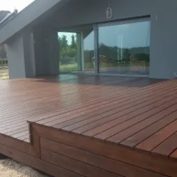 Nowoczesny taras z ciemnego drewna, z widoczną konstrukcją nośną i dużymi przesuwnymi drzwiami balkonowymi w tle, z widokiem na wnętrze domu.