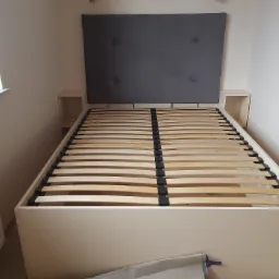 Rama łóżka z drewnianym stelażem i szarym zagłówkiem, z wbudowanymi szafkami nocnymi po bokach, ustawiona w rogu pokoju obok grzejnika.