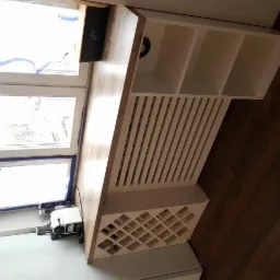 Drewniany blat kuchenny przy oknie z osłoną grzejnika z białymi lamelami i półkami, nad białą lodówką, widok z boku.