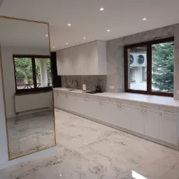 Nowoczesna kuchnia z białymi szafkami, złotymi uchwytami i marmurowym wykończeniem ściany oraz podłogi, widok z lustra w złotej ramie.