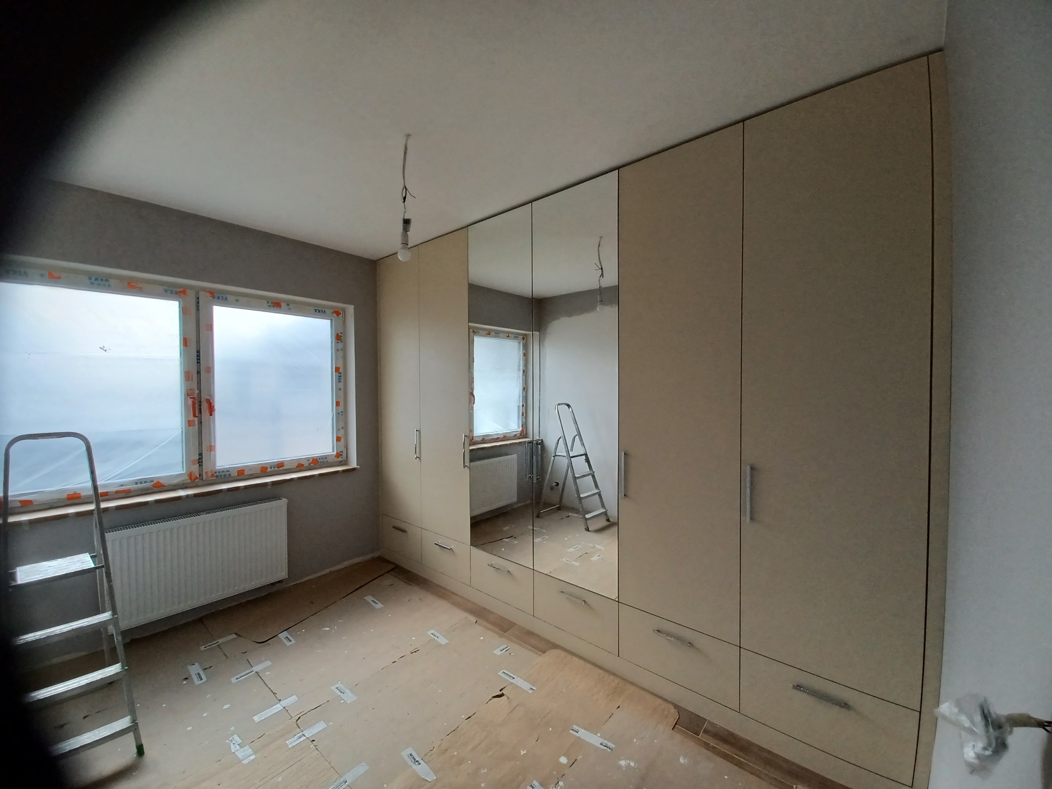 Beżowa szafa wnękowa z lustrzanymi drzwiami w trakcie montażu, widoczne zabezpieczone okno, drabina i ślady po montażu na podłodze.
