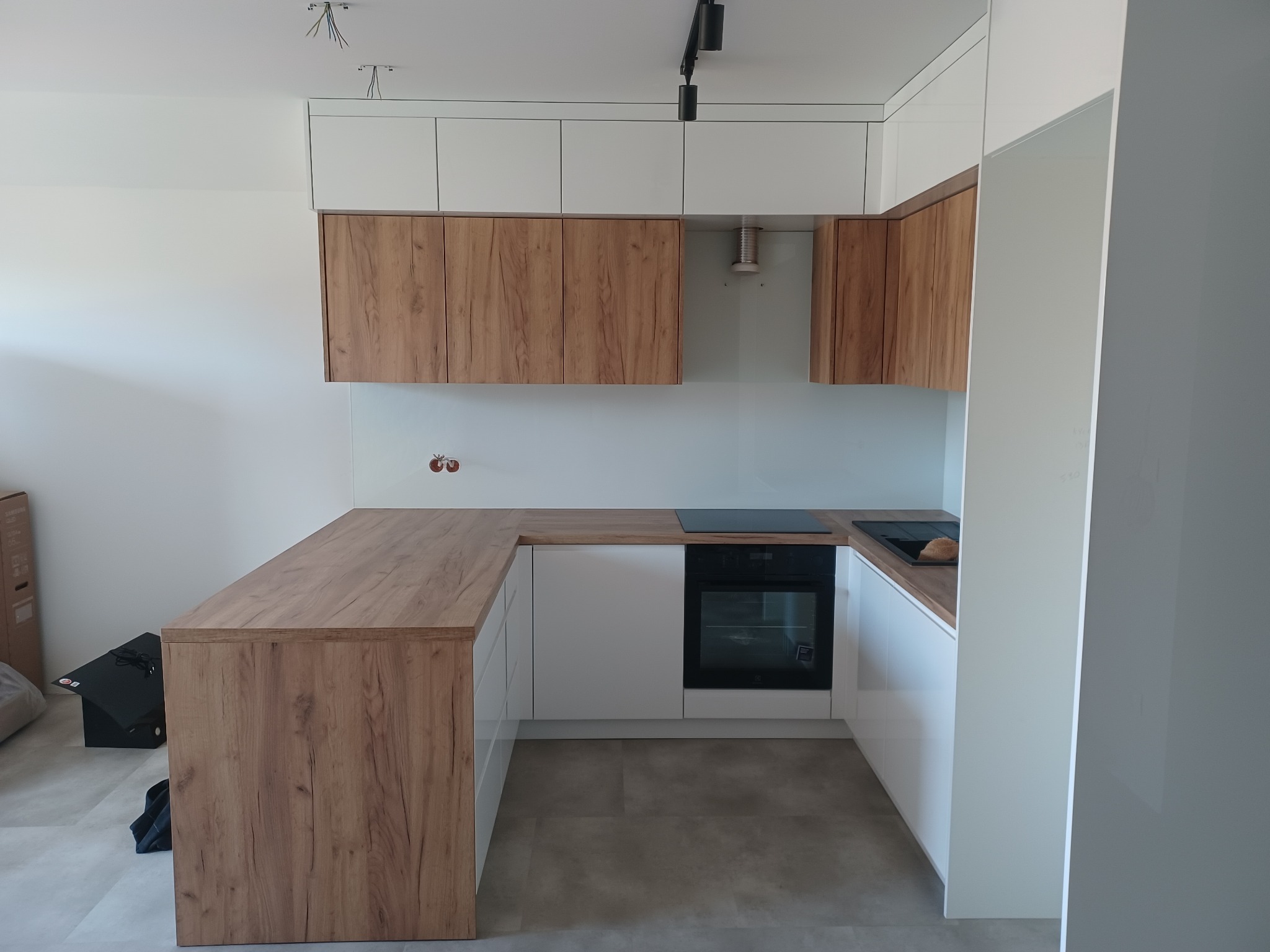 Nowoczesna kuchnia w kształcie litery U z drewnianymi szafkami wiszącymi i białymi szafkami dolnymi, czarnym piekarnikiem i płytą grzewczą, oraz szklanym panelem nad blatem.