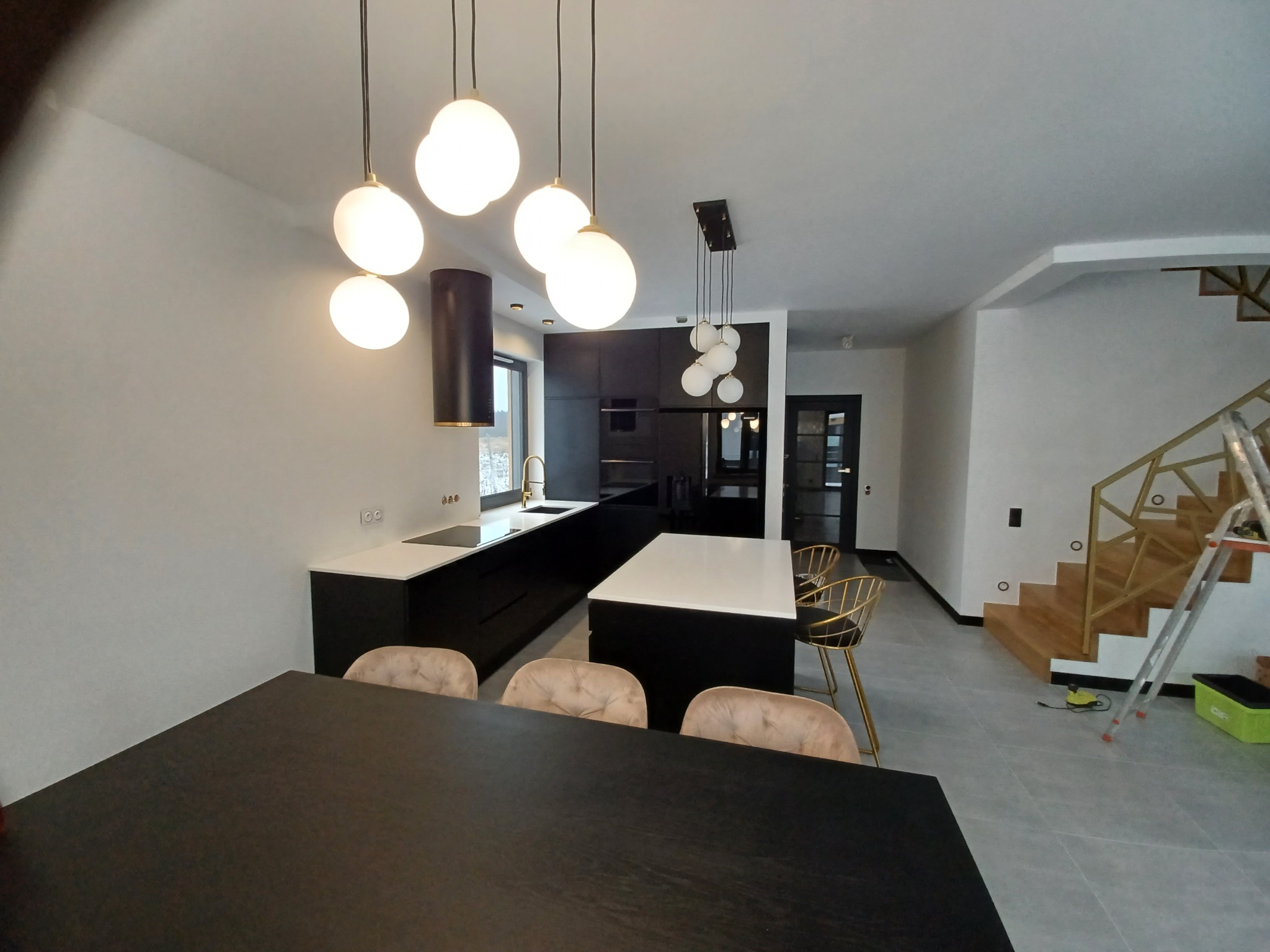 Nowoczesna kuchnia w czerni i bieli z wyspą, oświetlona wiszącymi lampami kulami, widok zza stołu jadalnego, schody w tle.