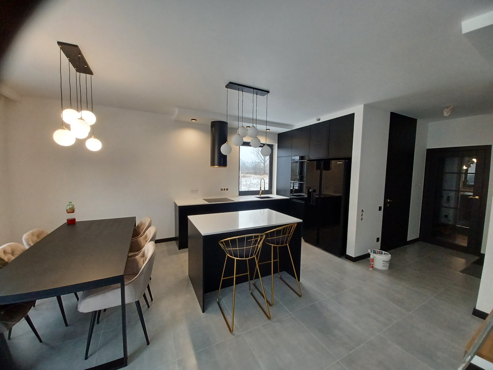 Nowoczesna kuchnia z czarnymi szafkami, białą wyspą, złotymi hokerami, szarymi płytkami podłogowymi i designerskim oświetleniem wiszącym.