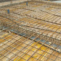 Zbrojenie płyty fundamentowej z widocznymi dystansami z tworzywa sztucznego, przygotowane do zalania betonem, w tle krajobraz z drzewami.