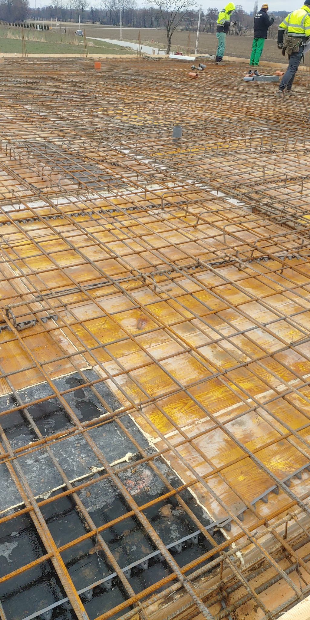 Siatka zbrojeniowa ułożona na szalunku z płyt OSB, przygotowana do zalania betonem, widoczne elementy dystansowe i pracownicy w tle.
