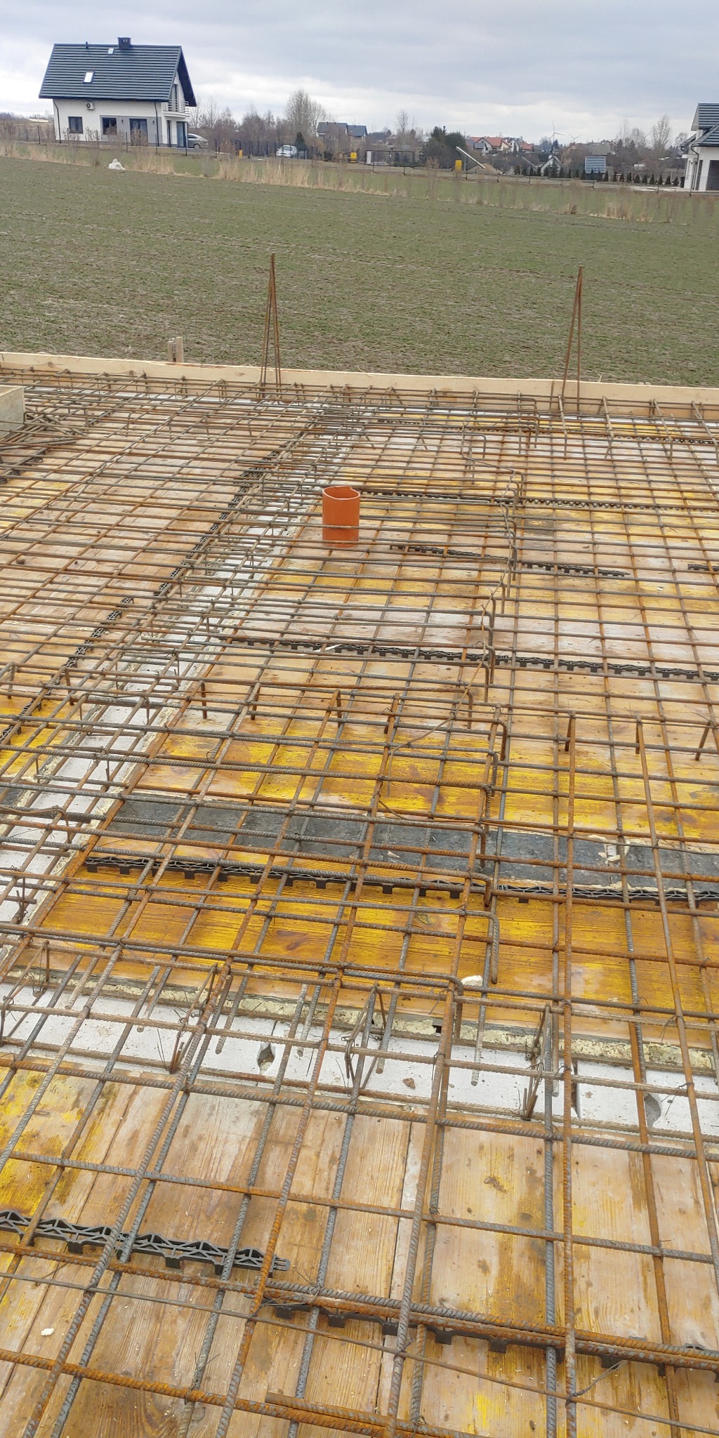 Zbrojenie stropu na deskowaniu z widoczną rurą wentylacyjną, w tle budynek mieszkalny i pole pod pochmurnym niebem.