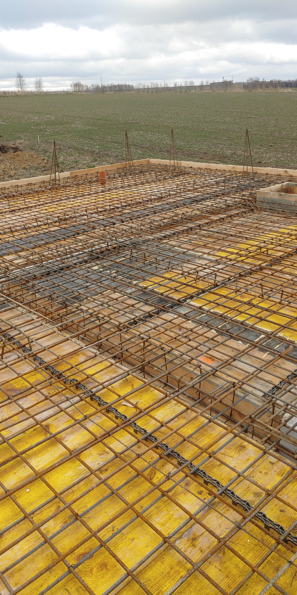 Zbrojenie fundamentu na żółtym szalunku z widocznymi dystansami z tworzywa sztucznego, w tle pole i pochmurne niebo.