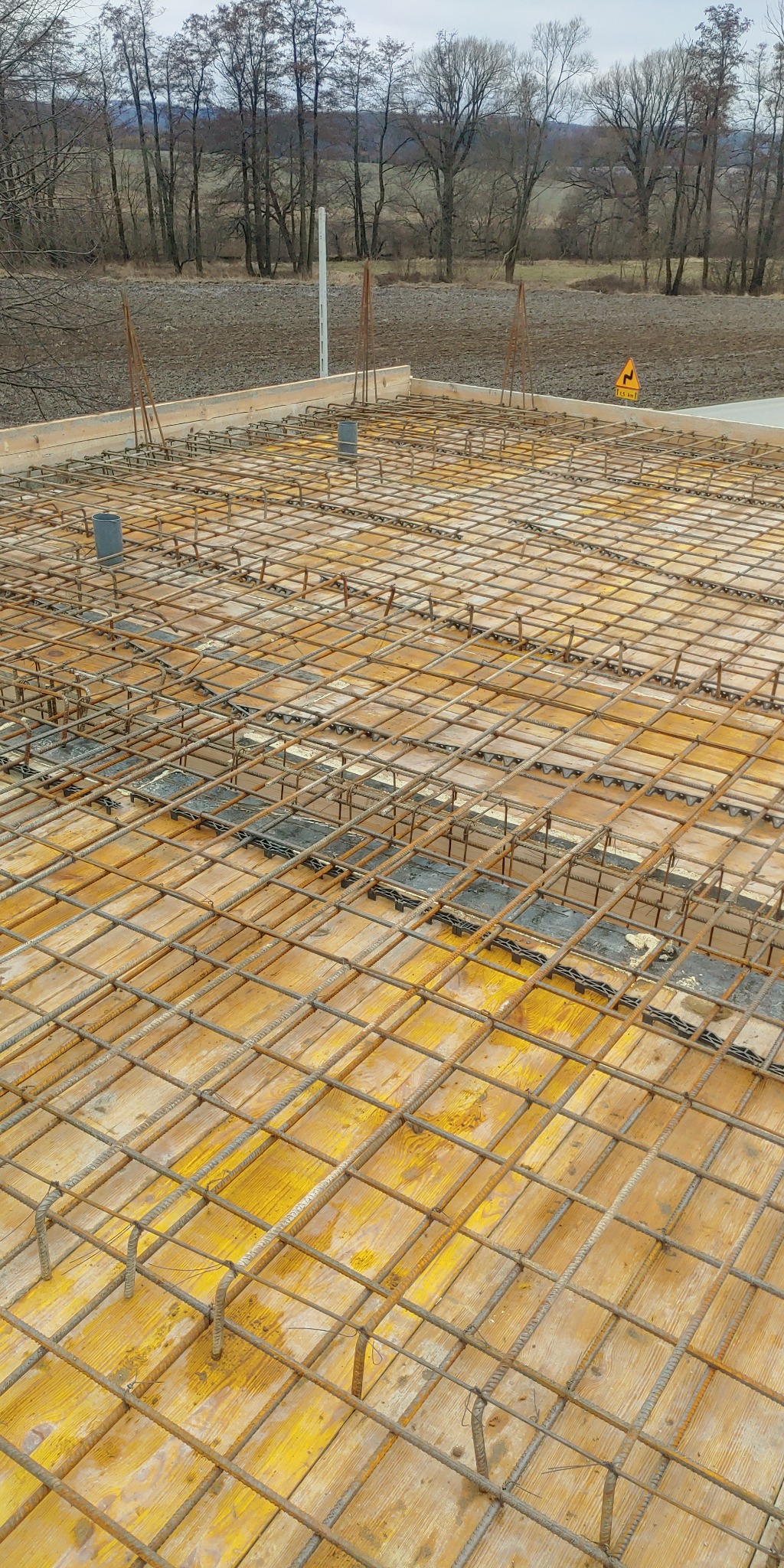 Zbrojenie płyty fundamentowej z widocznymi dystansami z tworzywa sztucznego, przygotowane do zalania betonem, w tle krajobraz z drzewami.