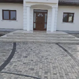 Nowoczesny podjazd z kostki brukowej w odcieniach szarości, prowadzący do wejścia domu z granitowymi schodami i kolumnami.