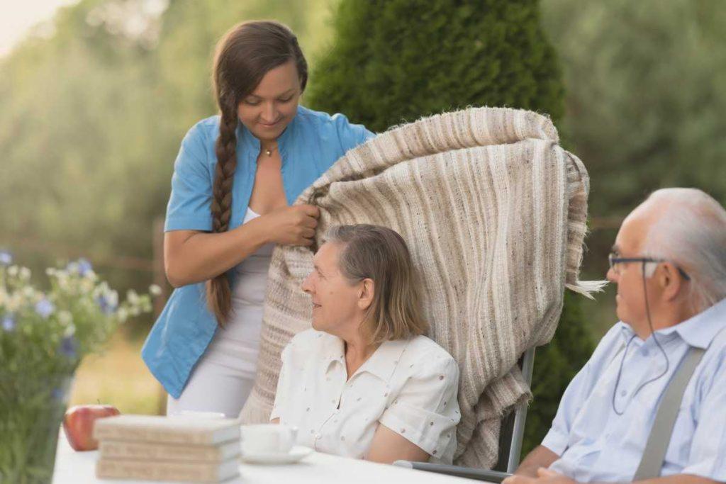 Opiekunka okrywa kocem starszą kobietę siedzącą przy stole na zewnątrz, obok siedzi starszy mężczyzna w okularach, w tle zieleń ogrodu.