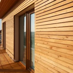 Drewniana elewacja domu z widocznymi poziomymi deskami, dużym oknem przesuwnym i drewnianym tarasem. Widok z zewnątrz, ciepłe oświetlenie.