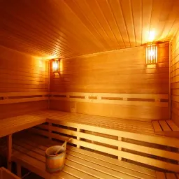 Wnętrze sauny wykończone drewnem, z ławkami, oświetleniem i drewnianym wiadrem. Ciepłe światło podkreśla naturalny kolor drewna.