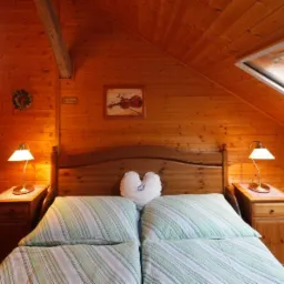 Drewniana sypialnia na poddaszu z łóżkiem, szafkami nocnymi i lampkami. Ściany i sufit wyłożone boazerią, okno dachowe. W tle widoczny grzejnik i dekoracje ścienne.