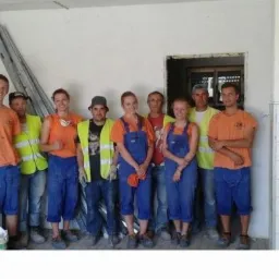 Grupa ośmiu osób, w tym kobiety i mężczyźni w roboczych ubraniach, pozuje w pomieszczeniu w trakcie prac wykończeniowych, widoczna drabina oparta o ścianę i częściowo wykończona ściana z białymi...