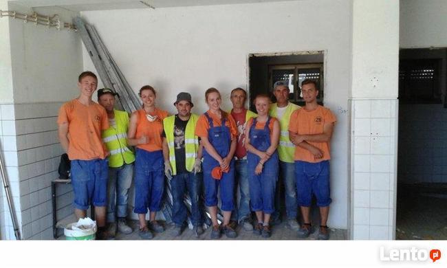 Grupa ośmiu osób, w tym kobiety i mężczyźni w roboczych ubraniach, pozuje w pomieszczeniu w trakcie prac wykończeniowych, widoczna drabina oparta o ścianę i częściowo wykończona ściana z białymi...