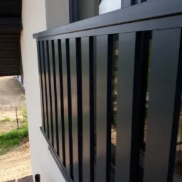 Nowoczesna, czarna, metalowa barierka balkonowa z pionowymi szczeblami, widok z góry na plac budowy i zieleń w tle.