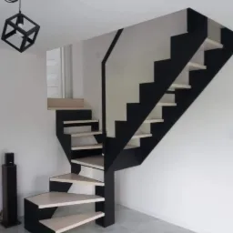 Nowoczesne schody z czarną konstrukcją i jasnymi stopniami, minimalistyczne oświetlenie w kształcie sześcianu, widoczne głośniki kolumnowe w tle.