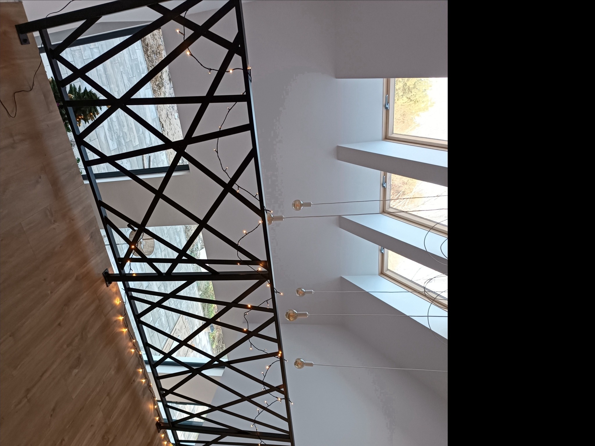 Wnętrze domu z czarną, geometryczną konstrukcją zabezpieczającą schody, udekorowaną lampkami, widok z dołu na okna dachowe i minimalistyczne lampy wiszące.