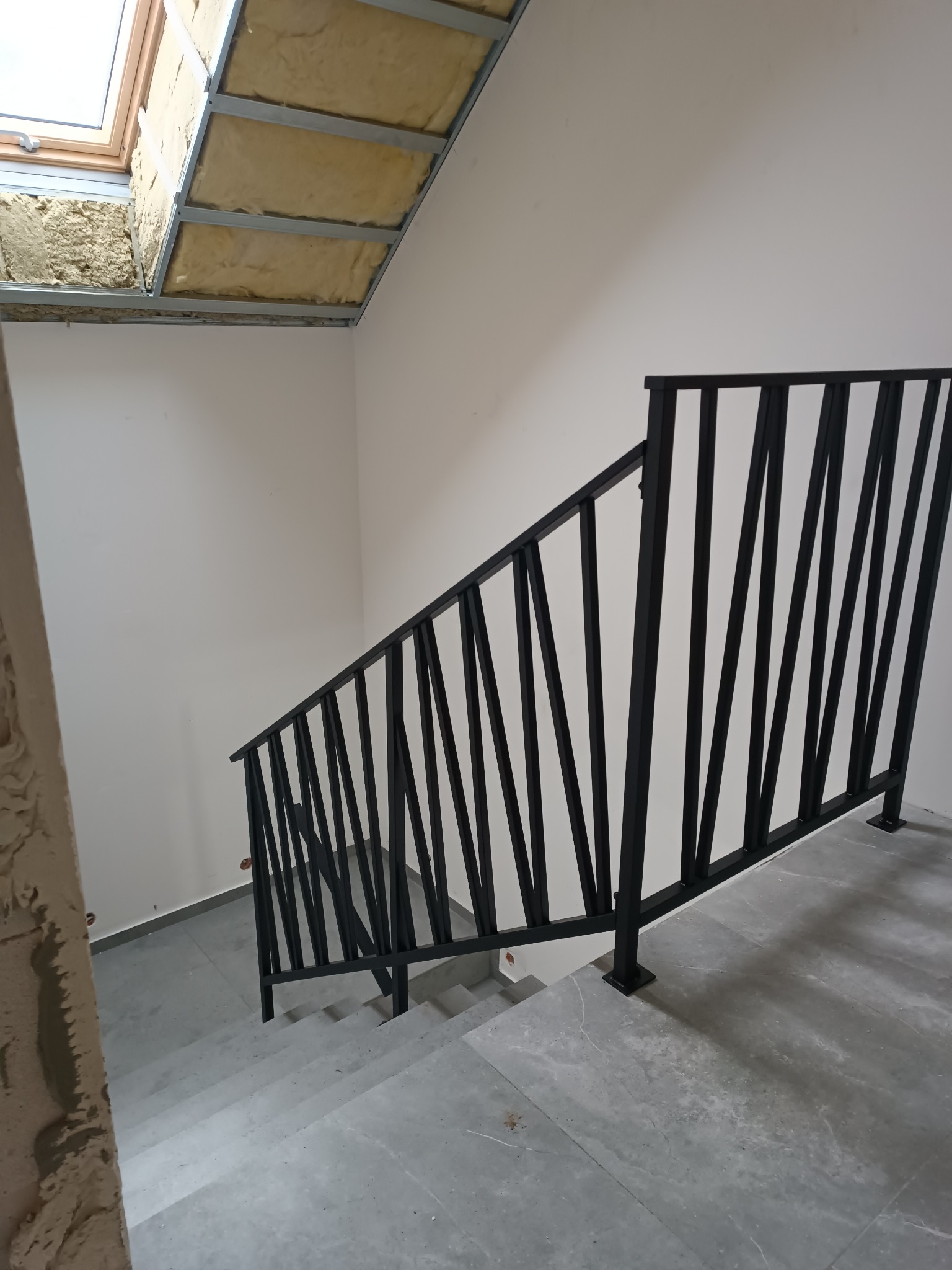 Nowoczesna, czarna barierka schodowa z pionowymi elementami o nieregularnym rozmieszczeniu, zamontowana w jasnym wnętrzu z szarymi płytkami podłogowymi i widocznym oknem dachowym w trakcie...