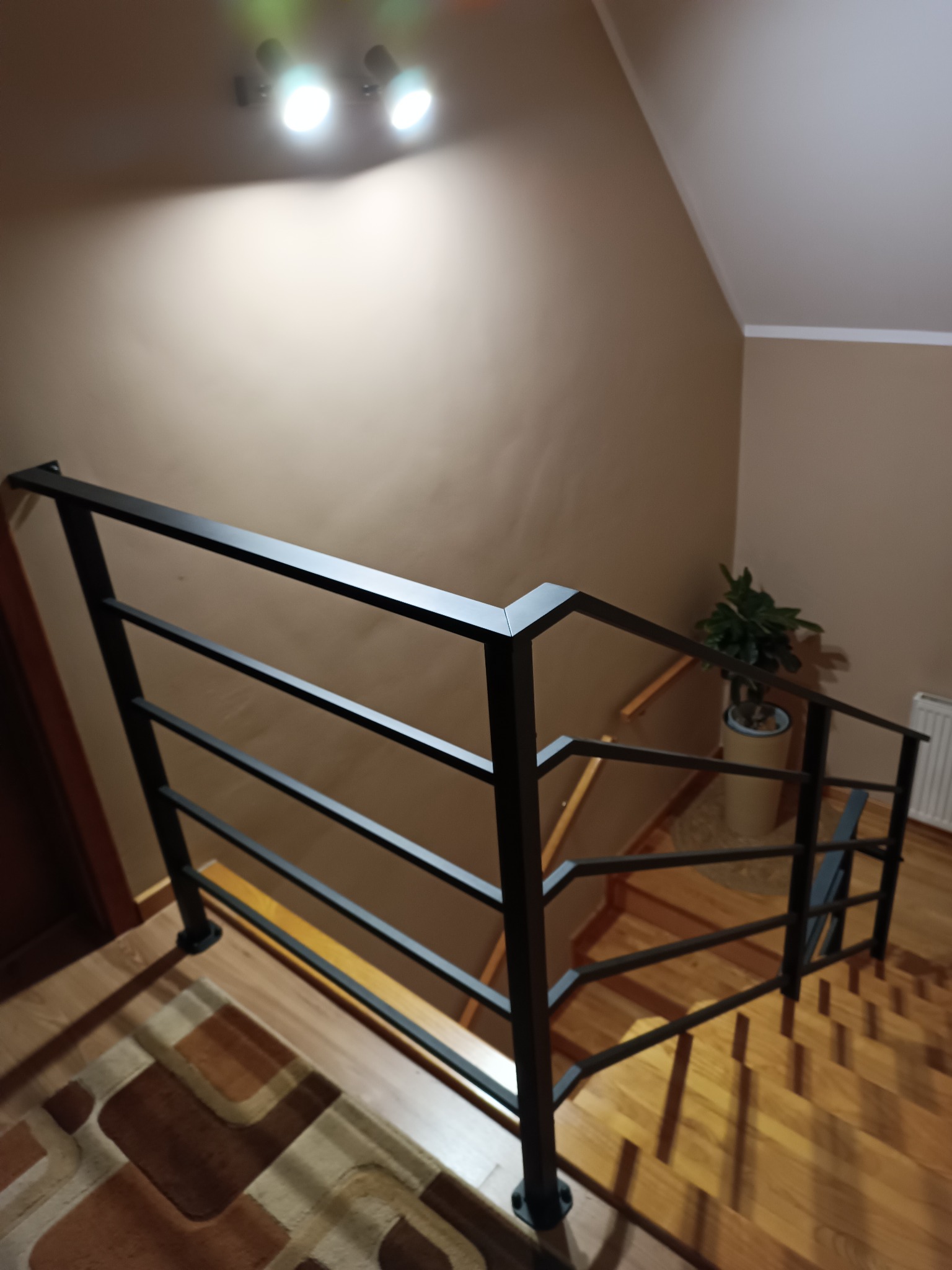 Schody z drewnianymi stopniami i nowoczesną, czarną, metalową barierką z poziomymi prętami, widok z góry, oświetlenie sufitowe, roślina doniczkowa w tle.