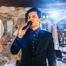 Wojciech Matkowski - Uśmiechnięty konferansjer w muszce i garniturze, trzymający mikrofon na tle kamiennej ściany i oświetlenia scenicznego. Widoczny sprzęt nagłośnieniowy.