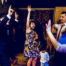 Wojciech Matkowski - Rozbawieni goście weselni tańczą na parkiecie, kobieta w kwiecistej sukience z uniesioną ręką, obok mężczyzna w rozpiętej koszuli i krawacie, w tle girlandy z chorągiewek.