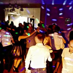 Wojciech Matkowski - Grupa osób tańczących w ciemnym pomieszczeniu oświetlonym kolorowymi światłami dyskotekowymi, widoczne wiszące lampy i fragment ściany z obrazem.