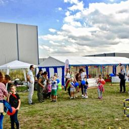 Wojciech Matkowski - Uroczystość plenerowa z białymi namiotami, dekoracjami i grupą dzieci z balonami na trawniku, w tle budynek przemysłowy i błękitne niebo z obłokami.