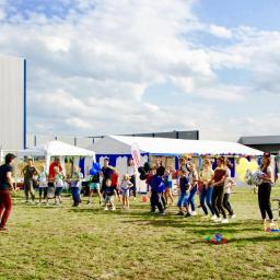 Wojciech Matkowski - Grupa dzieci i dorosłych bawi się na trawie podczas imprezy plenerowej, w tle białe namioty i budynki przemysłowe.