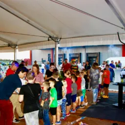 Dzieci ustawione w kolejce pod namiotem podczas imprezy plenerowej, w tle widoczny klaun i osoba z makijażem, prawdopodobnie animatorzy.