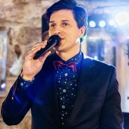 Wojciech Matkowski - Mężczyzna w garniturze i bordowej muszce śpiewa do mikrofonu na tle sceny z oświetleniem w Poznaniu.