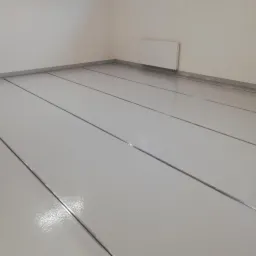 Jasnoszara, gładka podłoga z metalowymi listwami dylatacyjnymi w pustym pomieszczeniu z grzejnikiem na ścianie.