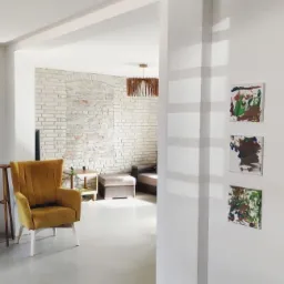 Jasne wnętrze z żółtym fotelem, białą cegłą na ścianie w tle i trzema abstrakcyjnymi obrazami dziecięcymi na białej ścianie z boku. Podłoga w odcieniach szarości.