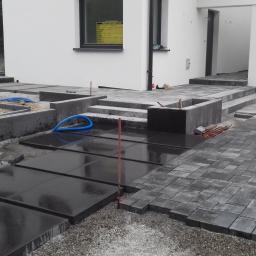 BolczasBruk - Montaż nawierzchni z kostki brukowej i płyt betonowych na posesji, widoczne przygotowane podłoże, częściowo ułożona kostka oraz płyty, węże do nawadniania i elementy konstrukcyjne.