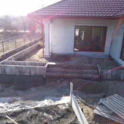 Przygotowanie terenu pod taras przy domu z widocznymi betonowymi elementami konstrukcyjnymi, podsypką żwirową i paletami budowlanymi, w tle budynek z brązową stolarką okienną i czerwoną dachówką.