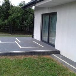 BolczasBruk - Nowoczesny taras z szarej kostki brukowej z białymi liniami tworzącymi geometryczny wzór, przylegający do białego budynku z dużymi przesuwnymi drzwiami.