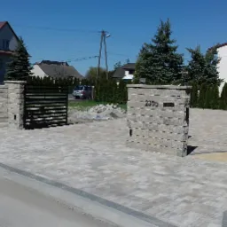 Ułożona kostka brukowa na podjeździe z czarną, metalową bramą i kamiennymi słupkami, widoczny numer domu 230a na jednym ze słupków.