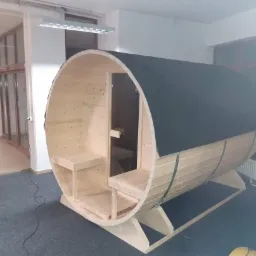 Surowa, drewniana sauna beczka z czarnym dachem, ustawiona w jasnym pomieszczeniu z wykładziną, widoczne wewnętrzne ławki i konstrukcja nośna.