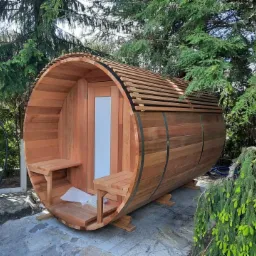 Drewniana beczka sauny ogrodowej z przeszklonymi drzwiami, umieszczona na betonowej powierzchni, otoczona zielenią drzew i krzewów.
